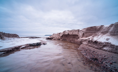 Tiny creek falls into Finnish Gulf though frozen sand dunes on Salmistu beach in Estonia. Empty coast in chilly winter morning, blue skyline over rural Nordic landscape and green conifer forest.