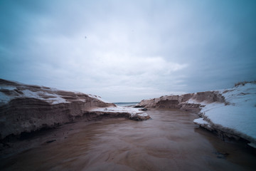 Tiny creek falls into Finnish Gulf though frozen sand dunes on Salmistu beach in Estonia. Empty coast in chilly winter morning, blue skyline over rural Nordic landscape and green conifer forest.