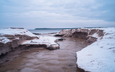 Tiny creek falls into Finnish Gulf though frozen sand dunes on Salmistu beach in Estonia. Empty coast in chilly winter morning, blue skyline over rural Nordic landscape and green conifer forest.