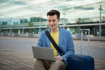 Stylish man sitting at airport with suitcase and laptop, working, typing, browsing. Businessman traveling. 