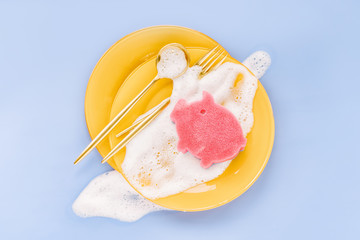 Yellow plates with spoons, forks and sponge on soapy foam background. Washing dishes concept. Flat lay, Top view.