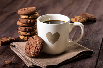 Cup of coffee with heart and chocolate chip cookies on rustic wooden background.