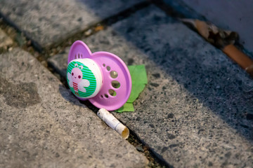 Dusseldorf, Germany - Grotesque Cigarette Butt Lying next to a Children Pacifier Nookie on a Street...