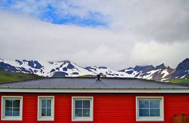 Red wooden house with snow capped mountains or glaciers on Iceland with blue sky and clouds