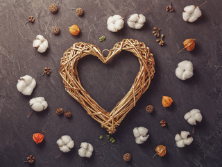 A golden wicker heart is on a black concrete background. Around are decorative cotton flowers and other dried plants. Romantic composition for Valentine's Day. Flat lay. Copy space.