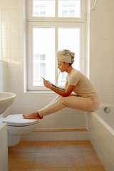 Beautiful young  girl in white towel sitting on a window in the bathroom looking at smartphone and brushes her teeth.