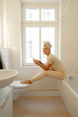 Beautiful young  girl in white towel sitting on a window in the bathroom looking at camera, holding smartphone and brushes her teeth.