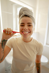 Pretty brown-eyed girl in white towel looks into camera and brushes her teeth in the bathroom.