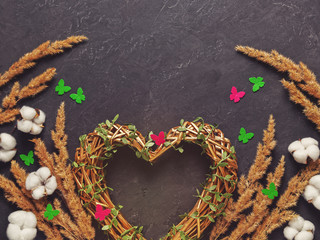 Romantic composition for Valentine's Day. A golden wicker heart is on a dark background. Decorating with dried plants, cotton flowers and colorful felt butterflies. Flat lay. Copyspace.