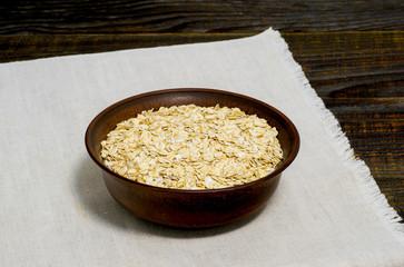 Oatmeal flakes in a ceramic brown bowl on a light linen napkin on a dark wooden table background.