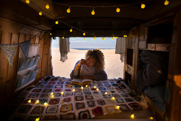 Travel and independent lifestyle concept with young beautiful curly free lady reading a book outside her hand made vintage wooden van and sandy sunny beach outdoor in background