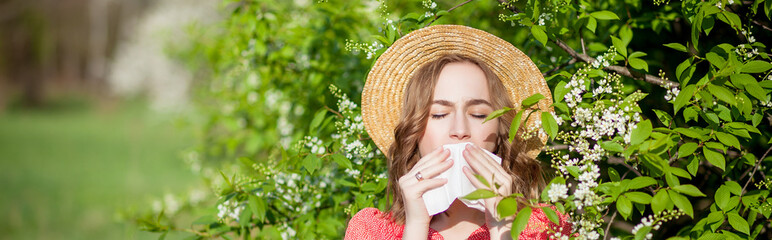 Young girl blowing nose and sneezing in tissue in front of blooming tree. Seasonal allergens...