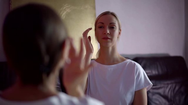 A close-up portrait of a blonde girl who is sitting opposite another girl is undergoing hypnosis practice in the room.