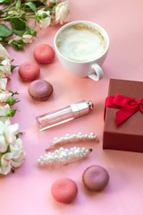 Flatlay pink coral background, the cup of cappuccino coffee and sweets macaroons, spring white roses, giftbox, beauty stuff - hair pin, nail polish and parfum. Best gift for woman