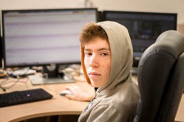 A portrait of a young guy in a hood is in front of the monitors. A teenager with facial pricks sits...