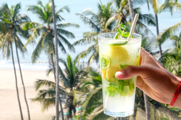 A young Asian mans hand holding a virgin mojito cocktail with palm trees the sea and a sandy beach in the background, the sun is shining, horizontal format