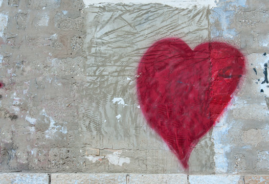 Red heart painted on the right side of a concrete old scratched grunge grey wall. A piece of a brick wall below. Copy space for text or images on the left hand side