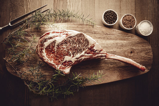 Raw dry aged wagyu tomahawk steak on a wooden background with salt, pepper, rosemary and a fork