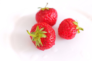 Three ripe juicy strawberry berries on a white plate are lit by bright light in an interesting foreshortening, close-up