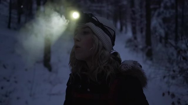 Slow motion of young woman breathing steam in headlamp alone in snow covered forest with river in winter