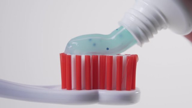 From a white tube, a man squeezes a blue toothpaste onto the red bristles of a plastic toothbrush. Personal hygiene products