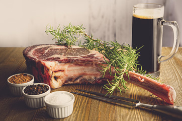 Raw dry aged wagyu tomahawk steak and a beer on a wooden background with salt, pepper, rosemary and a fork