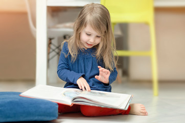 Little fair-haired girl examines illustrations in a children's book sitting on the floor at home....