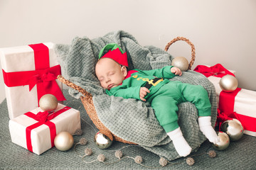 Little baby in costume of elf and with Christmas gifts sleeping in basket at home