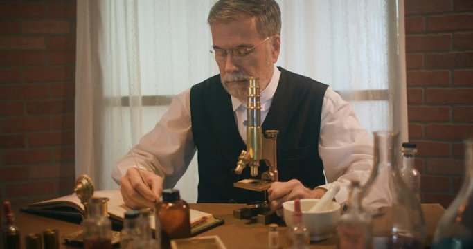 Scene tracks by a 1890 era scientist working in his laboratory using a dip pen to make some notations in a notebook of his work.