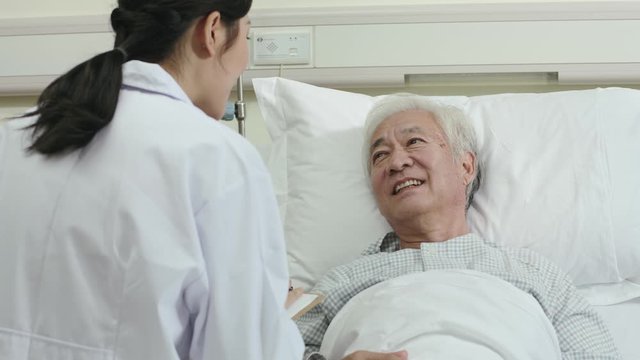 friendly young asian female doctor talking to male senior patient at bedside in hospital ward