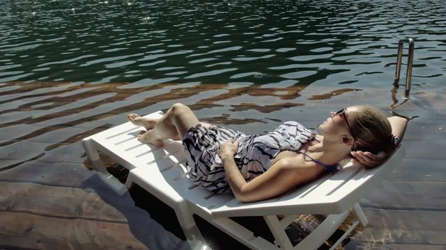 Woman lie on a sunbed in sunglasses and a boho silk shawl. Girl rest on a flood wood underwater pier. The pavement is covered with water in the lake. In the background are mountain and a forest.