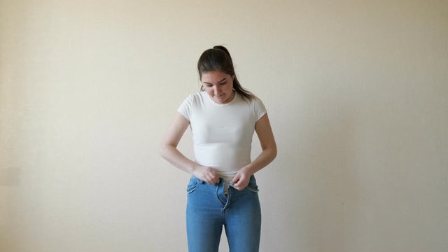 A woman in a white t-shirt bounces into her jeans and struggles to button them.