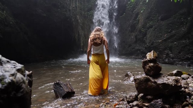 A slow motion shot of a young woman with yellow pants is walking between rocks, into a pool of water towards a small waterfall and then sits down. Pants are blowing in the wind from the waterfall.