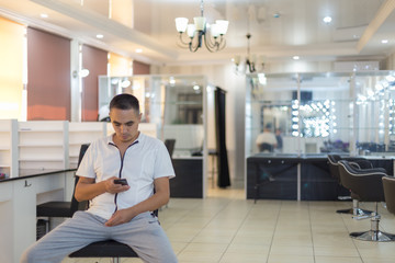 Naklejka na ściany i meble A man sits on a chair in the center of a beauty salon in the hands of a smartphone