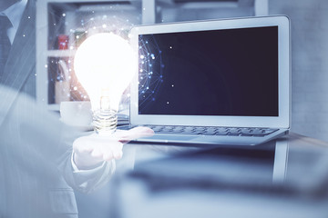 Computer on desktop in office with bulb icon hologram. Multi exposure. Concept of idea.