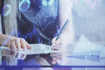 A woman hands writing information about stock market in notepad. Forex chart holograms in front. Concept of research.