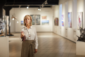 Portrait of mature female expert posing in modern art gallery hall looking at camera and holding clipboard, copy space