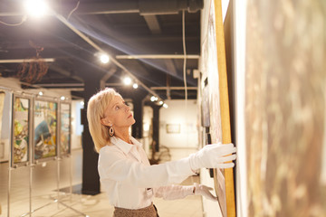 Wide angle portrait of elegant mature woman hanging paintings while working in art gallery or...
