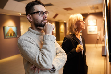 Waist up portrait of bearded man looking at paintings while enjoying exhibition in modern art gallery or museum, copy space