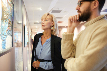Waist up portrait of mature couple looking at paintings while enjoying exhibition in art gallery or...