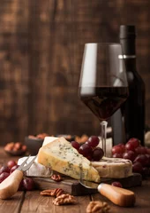  Glass and bottle of red wine with selection of various cheese on the board and grapes on wooden background. Blue Stilton, Red Leicester and Brie Cheese and bowl of nuts. © DenisMArt