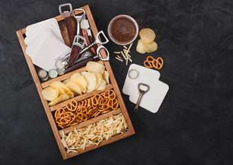 Glass of craft lager beer and opener with box of snacks on black background. Pretzel and crisps and salty potato sticks in vintage wooden box with openers and beer mats. Top view