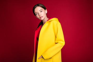 Serious calm and peaceful young woman look straight on camera in yellow hoody or coat. Stand alone. Fashionable clothes. Isolated over red background. Listen to music through wireless earphones.
