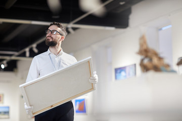 Waist up portrait of bearded art gallery worker holding painting while organizing exhibition in...
