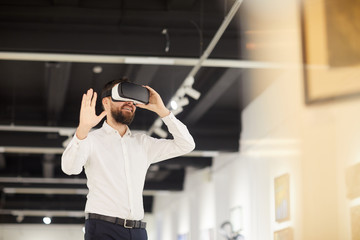 Waist up portrait of modern bearded man wearing VR gear while enjoying immersive experience during...