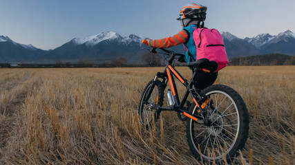 Obraz premium One caucasian children walk with bike in wheat field. Little girl walking black orange cycle on background of beautiful snowy mountains. Biker stand with backpack and helmet. Mountain bike hardtail.