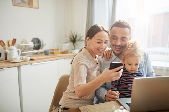 Warm-toned portrait of modern mixed race-family using computer devices while sitting in cozy kitchen interior with cute little daughter, copy space