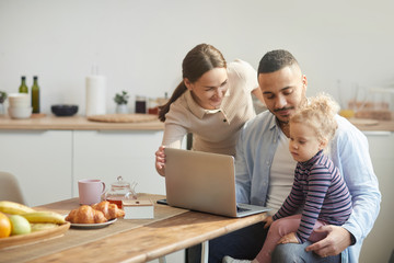 Warm-toned portrait of modern mixed race-family using computer while sitting in cozy kitchen...