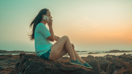 Relaxed woman sitting on stone and speaking on smartphone on shore. Side view of pleasant woman spending time enjoying vacation and talking on mobile phone on stony shore