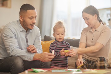 Warm toned portrait of happy mixed-race family playing with cute little girl in cozy home interior